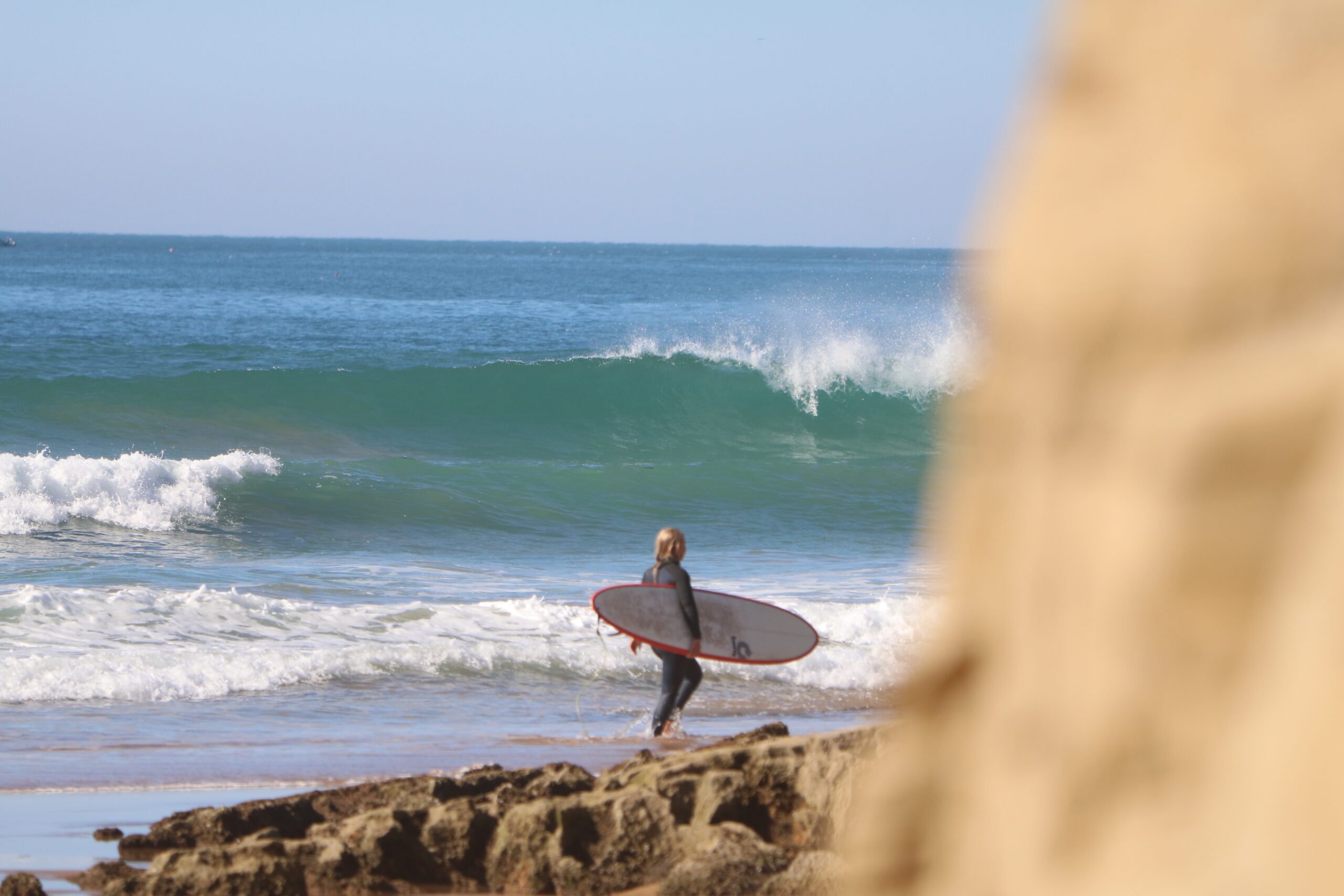 A girl holding a surfboard on Desert Point beach in Morocco, with a breaking wave in the background, for the article about surfing in Morocco in September.