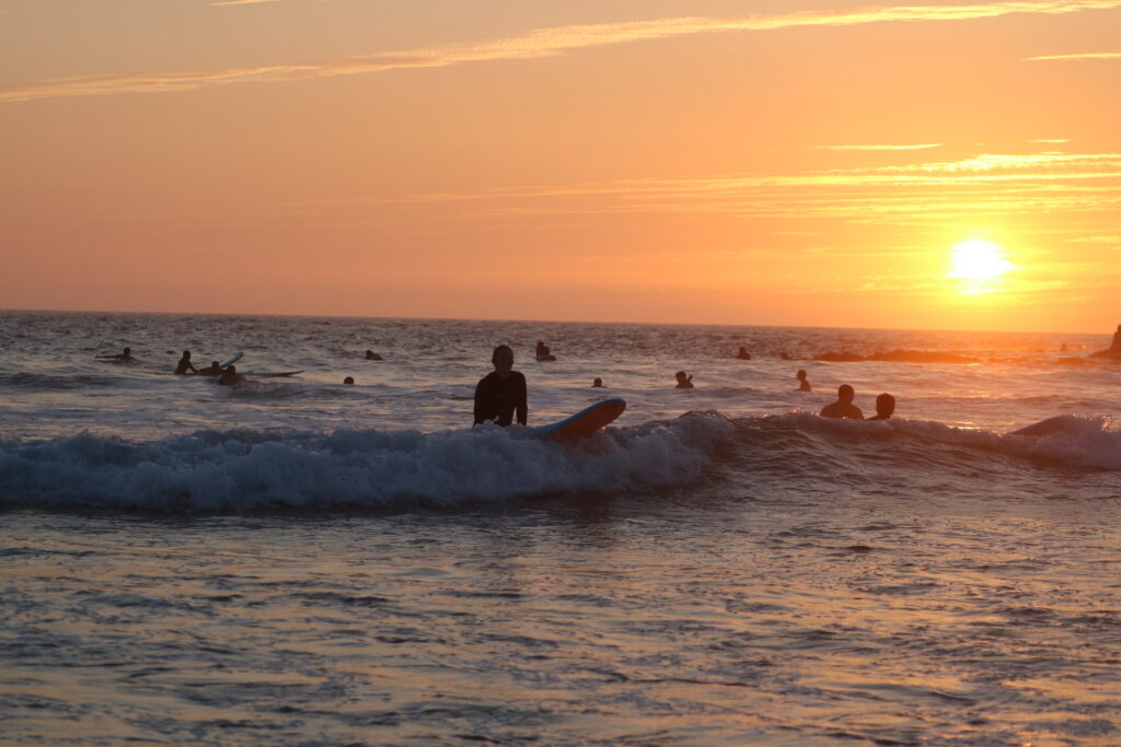 Un surfeur glissant sur une vague au coucher du soleil, entouré de couleurs dorées et orangées, capturant un moment paisible et emblématique d'une session de surf en fin de journée au Maroc.
