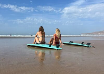 Dos chicas en la playa con sus tablas de surf al lado, listas para una sesión de surf en Marruecos, en un surf camp en Taghazout.